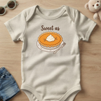 A baby short-sleeve bodysuit in a light oatmeal color, laid flat on a light wood surface. The bodysuit features a graphic of a pumpkin pie with whipped cream and the text 'Sweet as' above it.